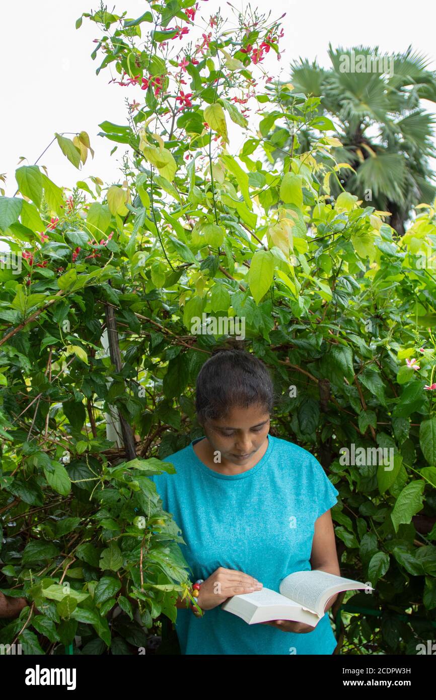 Una bella donna in cima blu legge un libro in luce del giorno, in piedi rilassato in un ambiente con piante verdi e fiori rossi sullo sfondo. Foto Stock