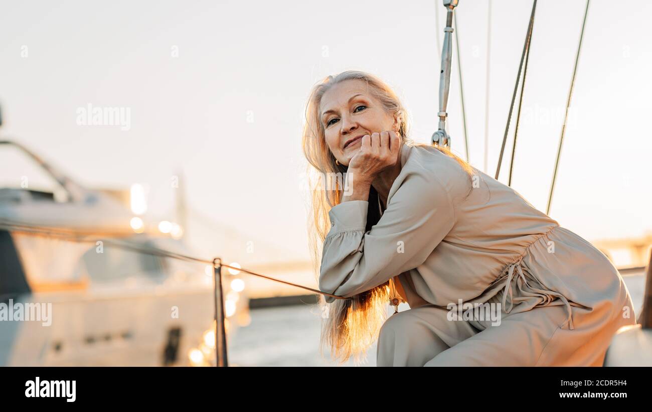 Ritratto di una donna caucasica matura con lunghi capelli grigi guardando direttamente alla macchina fotografica mentre si siede su una barca a vela a. tramonto Foto Stock