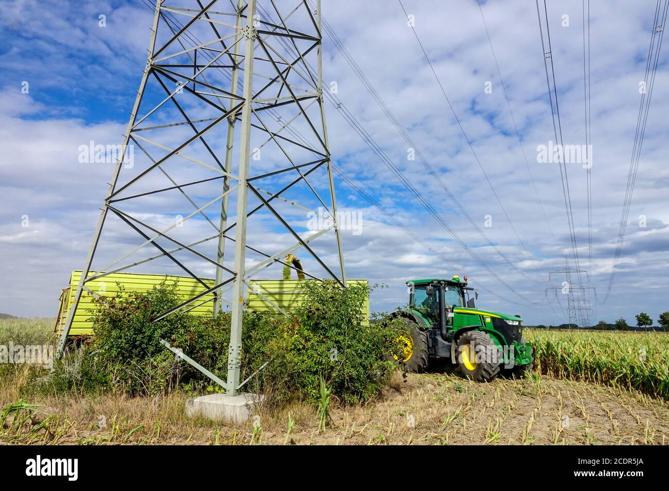 Agricoltura che raccoglie mais sotto linee ad alta tensione e piloni John Il trattore Deere aggira un montante Foto Stock