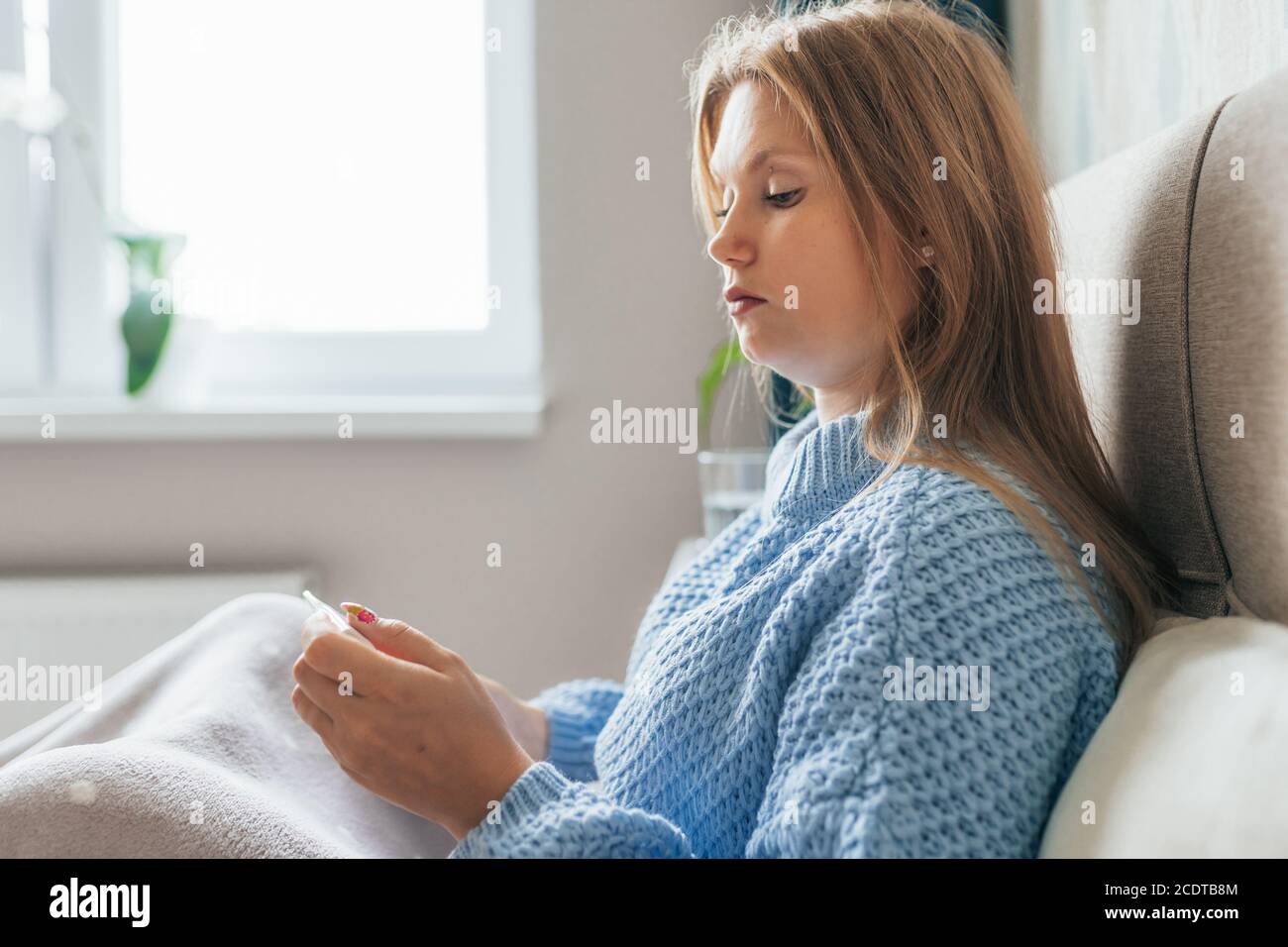 Malata giovane donna bionda si trova a letto a casa sotto coperta e controllare termometro. Mal di testa, influenza, freddo, virus. Foto Stock
