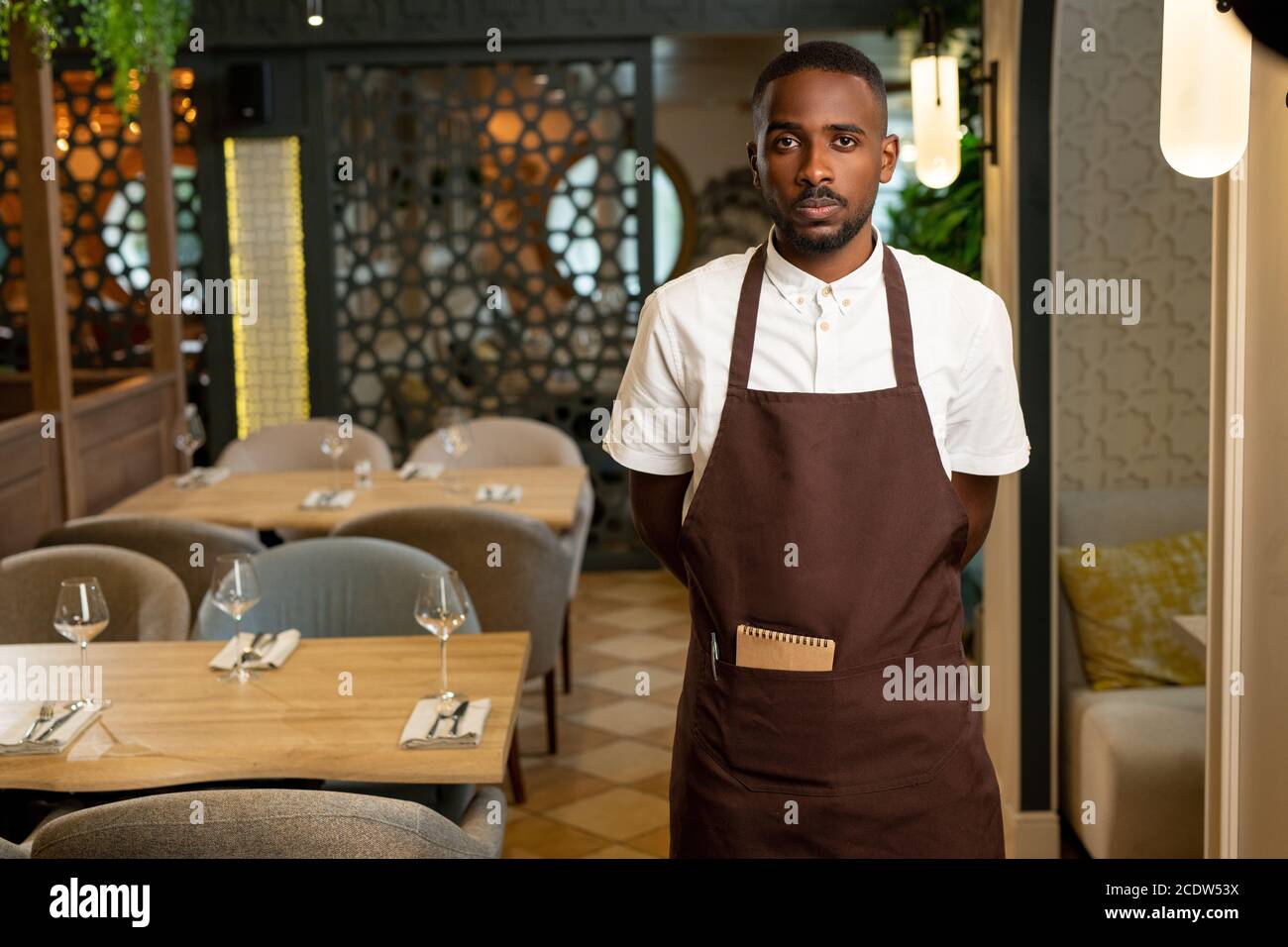 Giovane cameriere di etnia africana in piedi uniforme in navata tra le tabelle Foto Stock