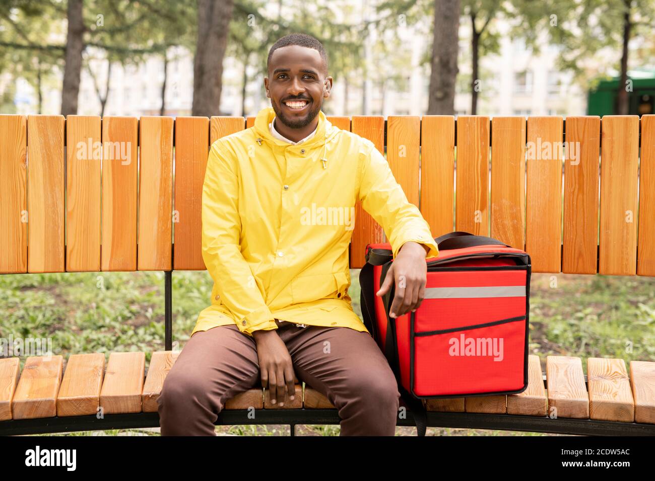 Giovane allegro corriere maschile in giacca gialla tenendo la mano sopra grande zaino rosso Foto Stock