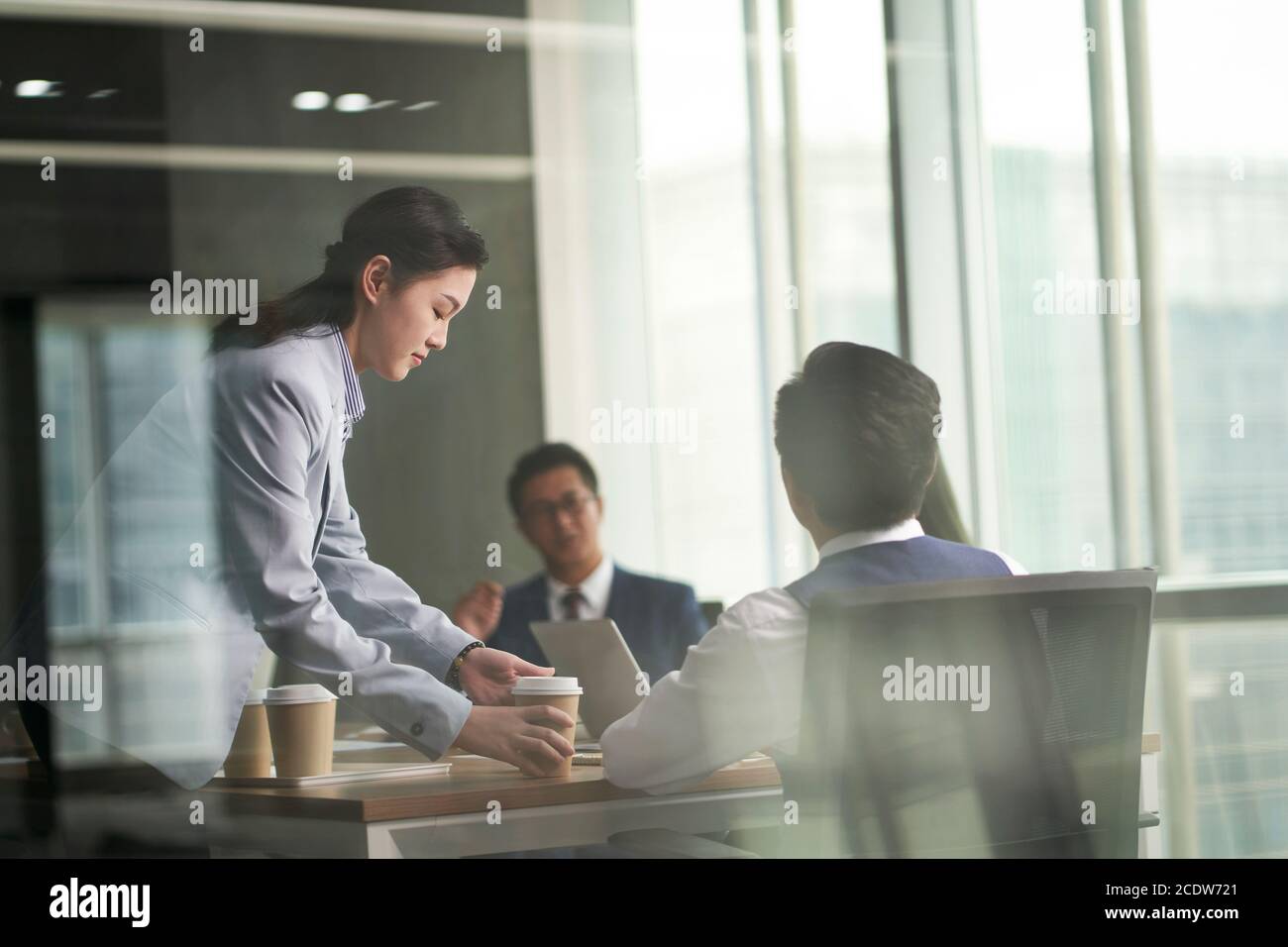 attraverso-il vetro colpo di un giovane intern asiatico tirocinante portando caffè alle persone d'affari che si riunano in ufficio Foto Stock