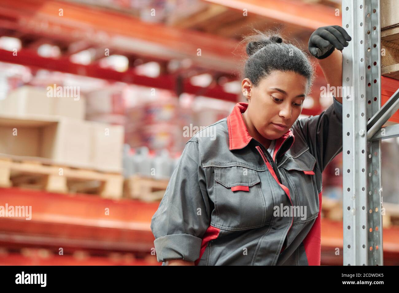 Giovane donna di razza mista stanca in uniforme e guanti appoggiati rack Foto Stock