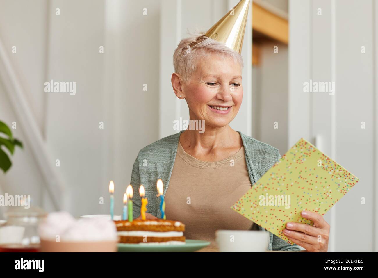 Donna anziana seduta al tavolo con lettura torta di compleanno un biglietto d'auguri e sorridente Foto Stock