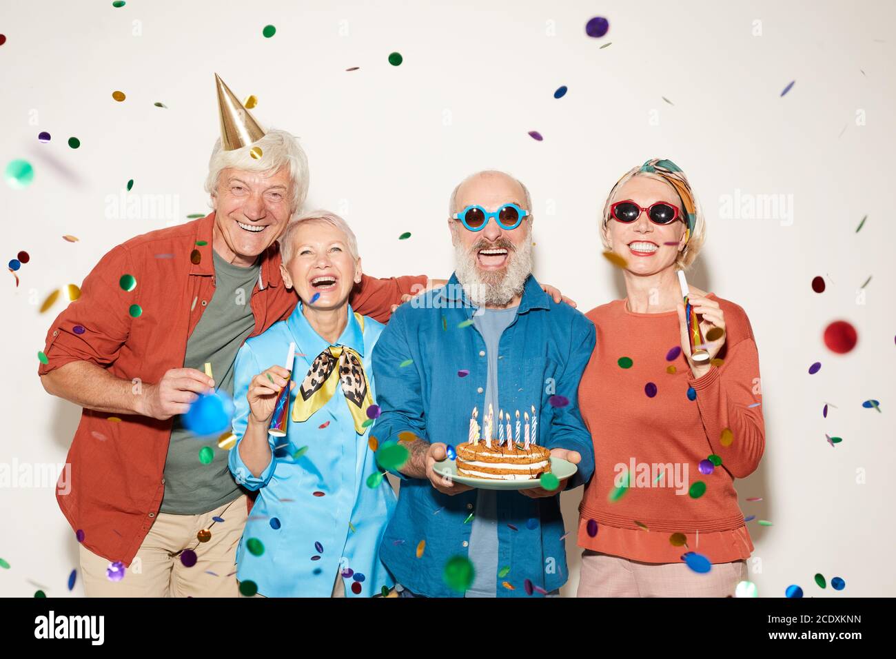 Ritratto di gruppo di anziani con torta di compleanno in piedi sotto i confetti e sorridendo a macchina fotografica contro il bianco sfondo Foto Stock