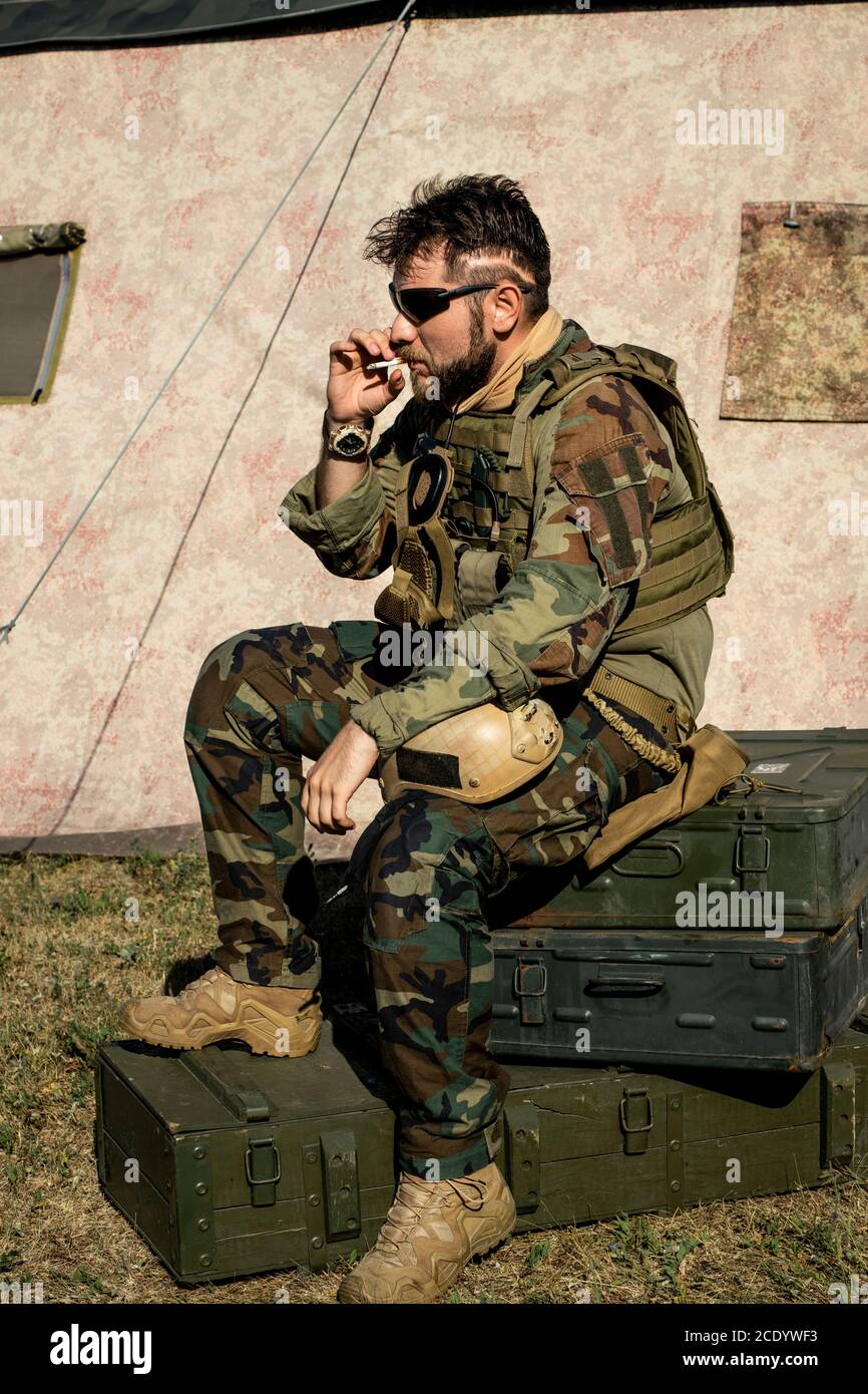 Uomo con portatore pensivo in abito camouflage seduto su valigie in metallo e fumare sigarette all'aperto Foto Stock