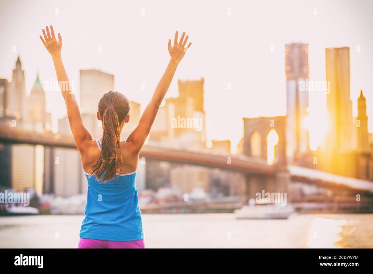 Donna felice in forma che raggiunge l'obiettivo di fitness a New York City con le braccia in su al tramonto e Ponte di Brooklyn. Ragazza corridore gioiosa ed eccitata dopo Foto Stock