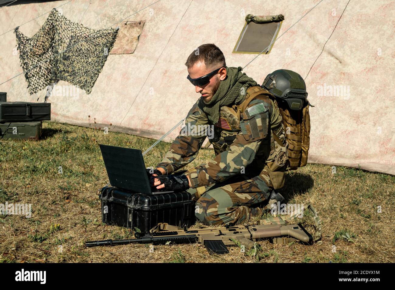 Concentrato giovane soldato programmatore seduto a terra e con cui lavora computer portatile durante l'analisi della rotta militare Foto Stock