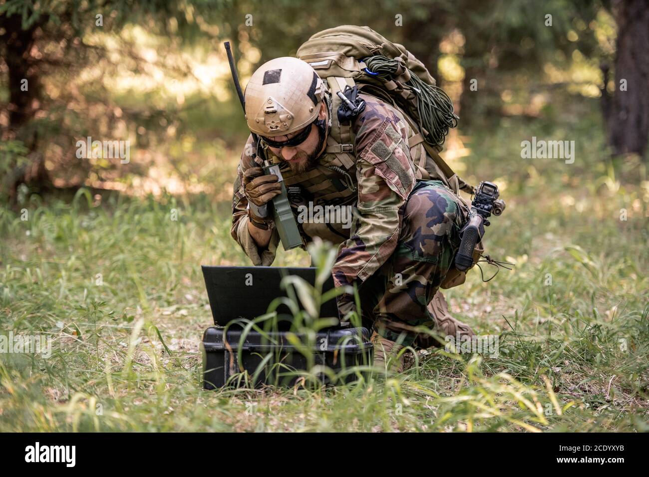 Soldato bearded in uniforme camouflage seduto in foresta e l'invio messaggio tramite dispositivo radio durante il controllo del funzionamento militare tramite computer Foto Stock