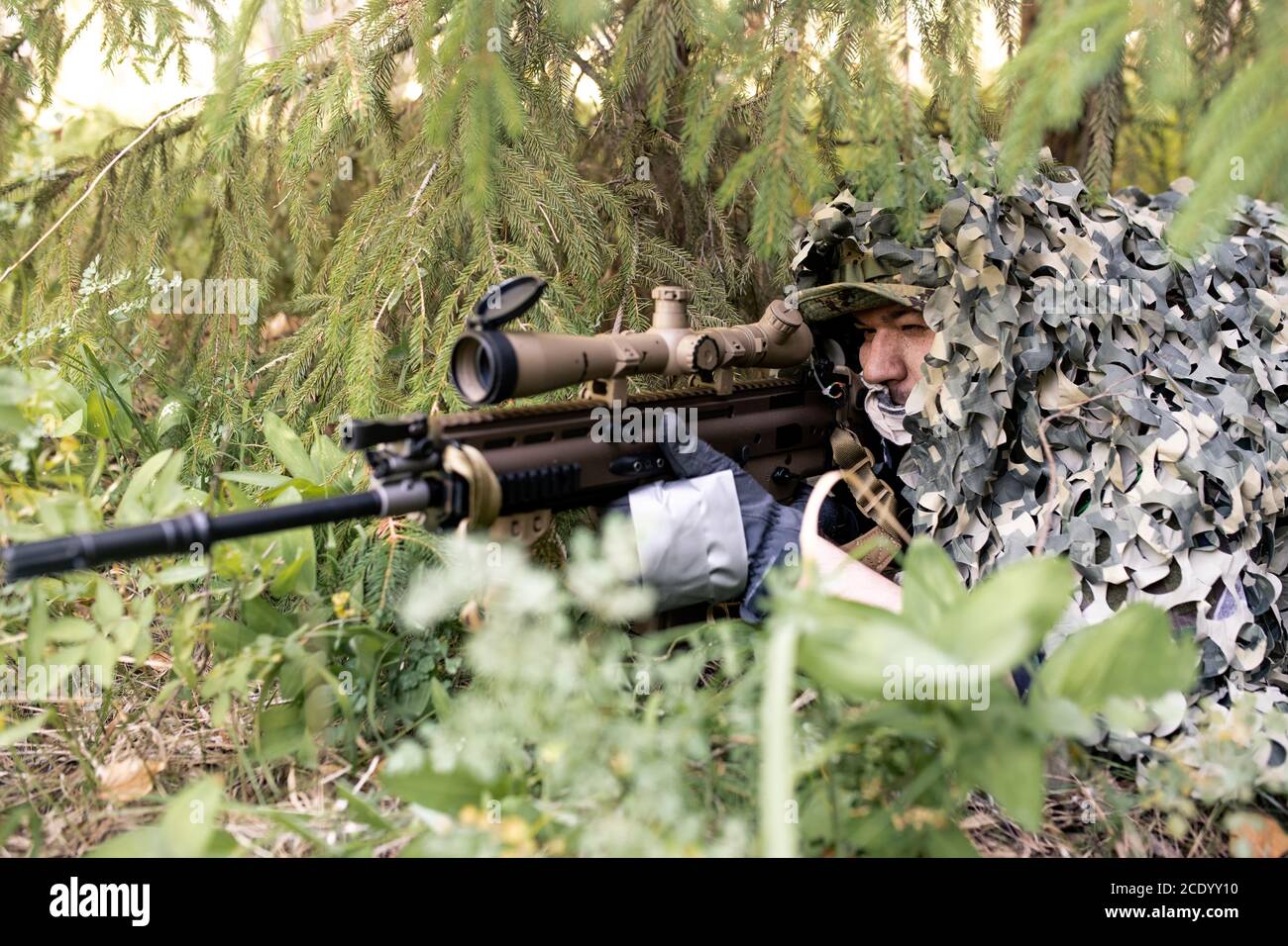 Cecchino concentrato che giace sotto rete di camuffamento su terra e guardando attraverso l'oscilloscopio del fucile Foto Stock