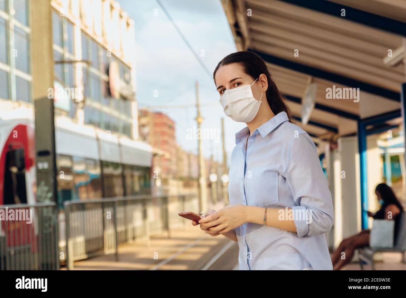 Foto di borsa di una donna che indossa una maschera facciale, tenendo un telefono e aspettando che il tram arrivi alla stazione. Nuovo concetto normale Foto Stock