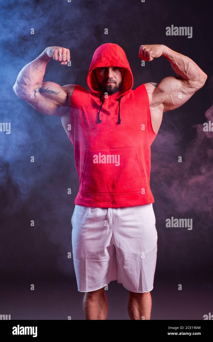 Potente uomo muscoloso con cappuccio in posa nella felpa rossa. Riprese in studio con fumo rosso e blu Foto Stock