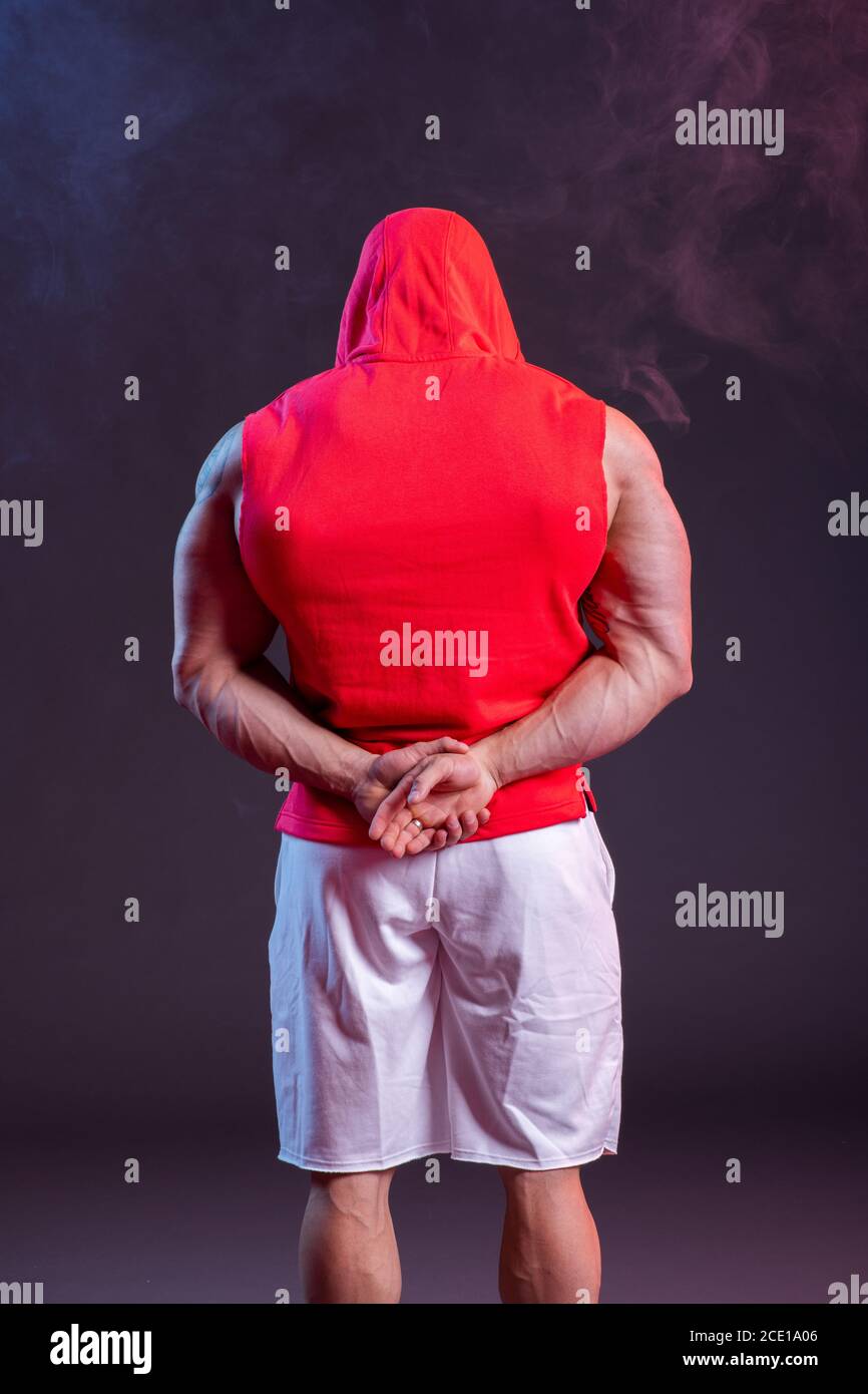 Potente uomo muscoloso con cappuccio in posa nella felpa rossa. Vista posteriore. Riprese in studio con fumo rosso e blu Foto Stock