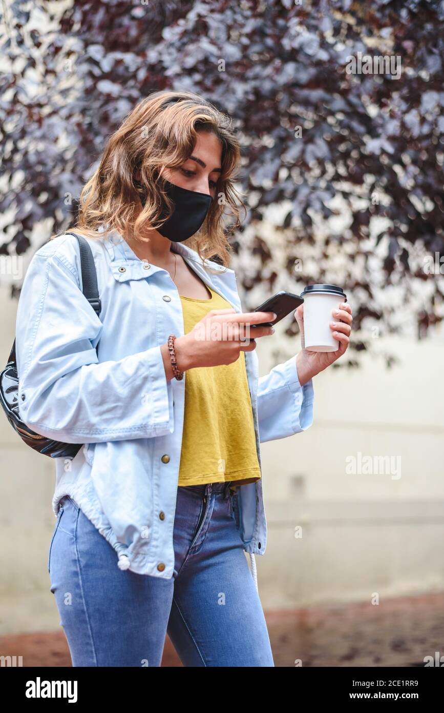 ritratto verticale di una giovane donna dai capelli rossi che indossa il marchio utilizzando telefono e tenendo una tazza di caffè da portare via dentro la strada Foto Stock