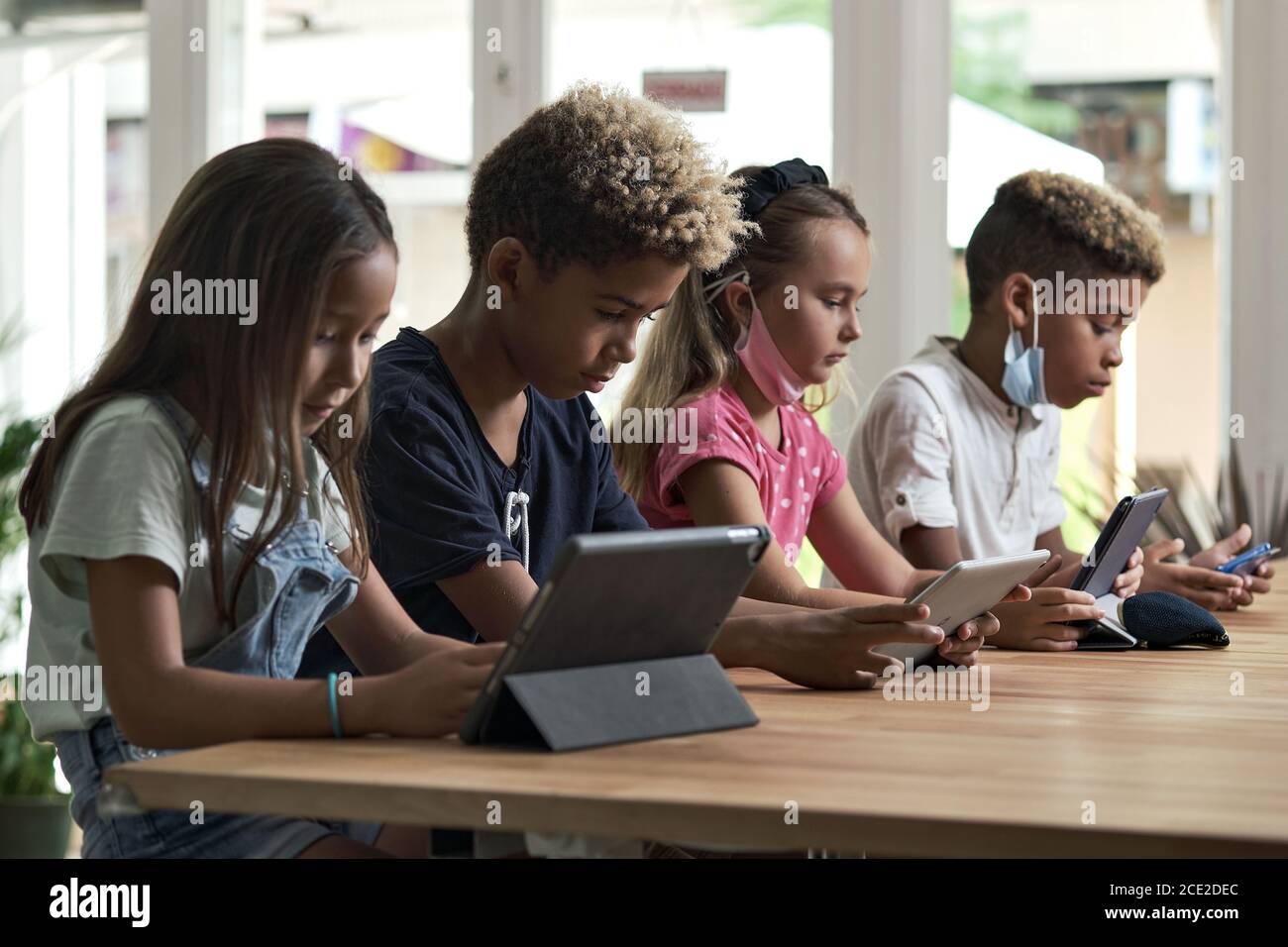 Quattro diversi bambini indossare maschere sedersi al tavolo utilizzare i gadget wireless ignorando l'uno l'altro preferiscono i giochi Internet e la comunicazione virtuale. Alfa generati Foto Stock