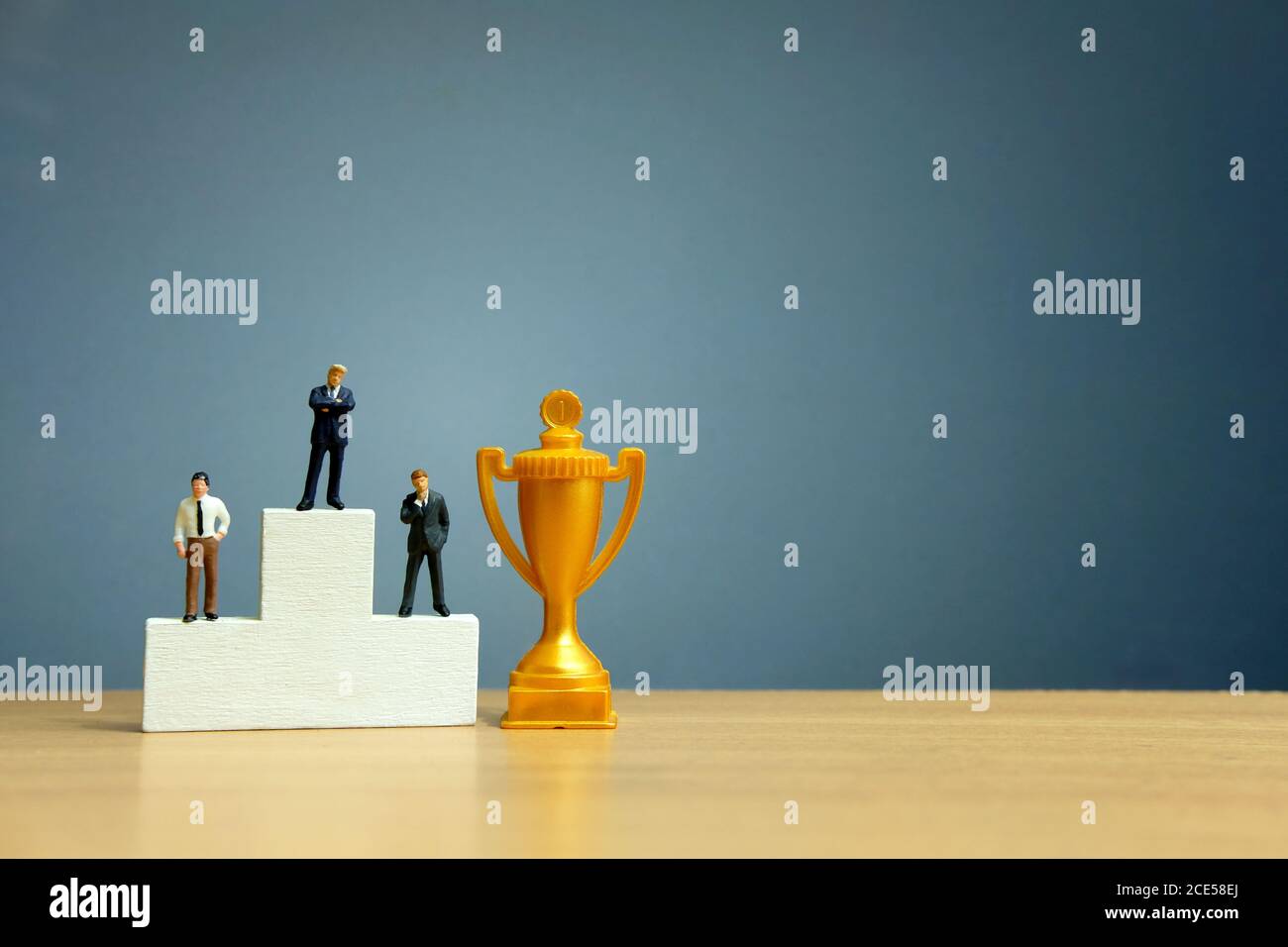Concetto di business in miniatura - uomo d'affari in piedi davanti al bianco podio vincitore con trofeo d'oro Foto Stock
