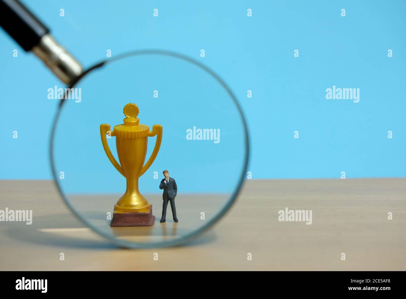 Concetto di business in miniatura - uomo d'affari in piedi di fronte all'oro trofeo e lente d'ingrandimento in vetro Foto Stock