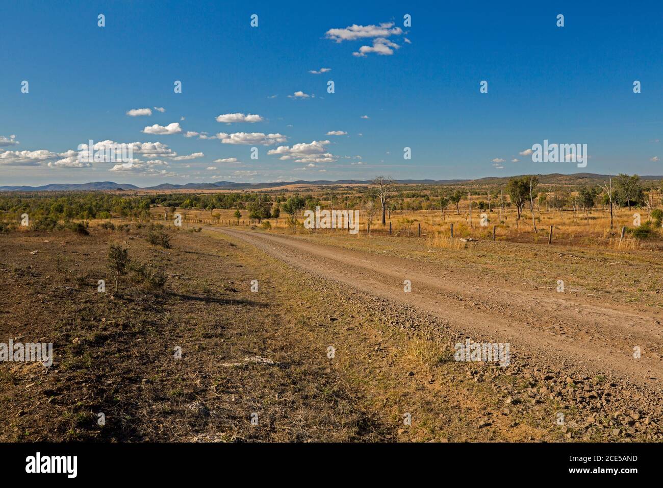 Arido paesaggio australiano dell'entroterra durante la siccità con la strada di ghiaia rossa spianando attraverso le pianure a basse catene montuose lontane sotto il cielo blu con le nuvole Foto Stock