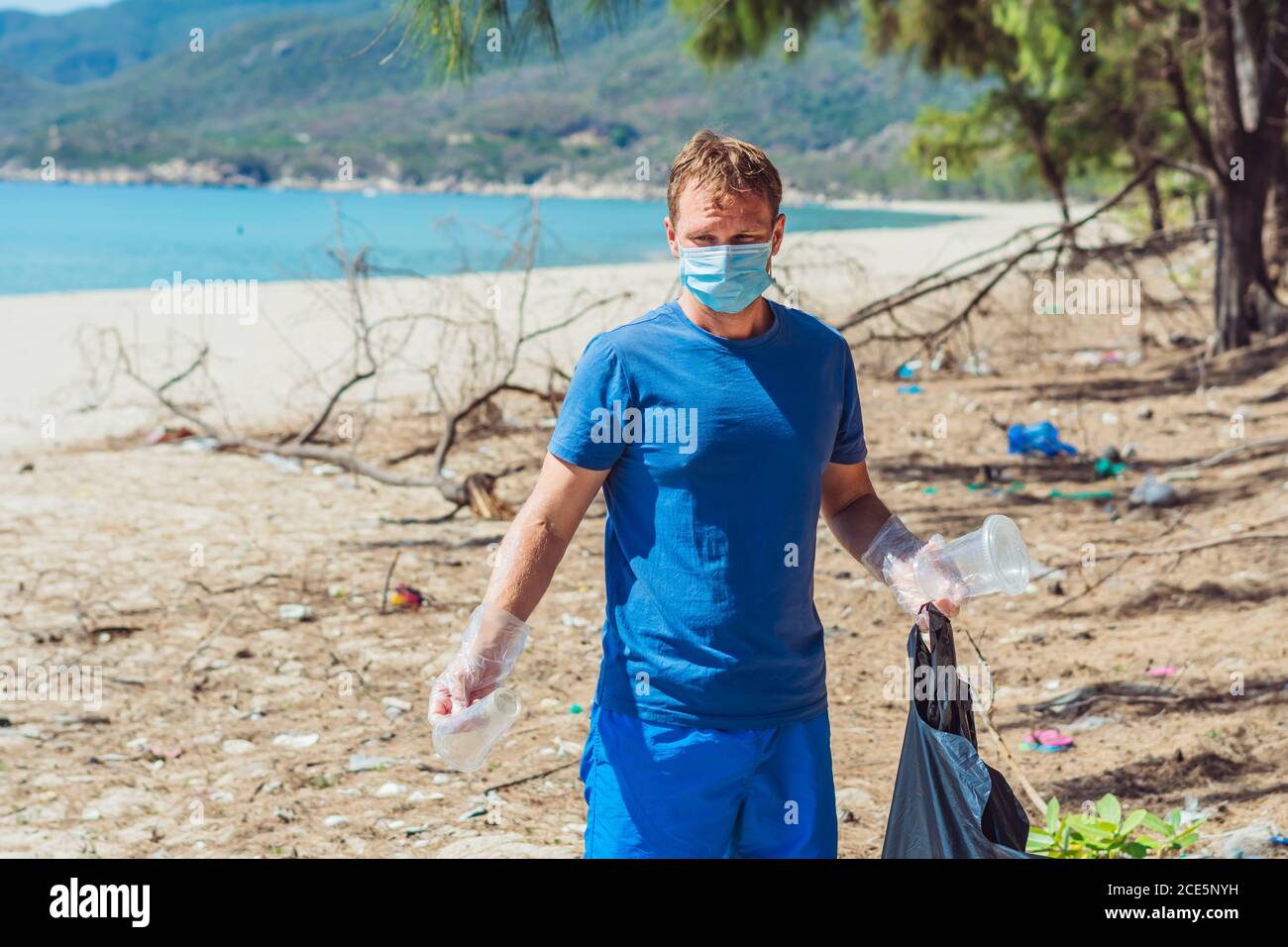 Volontario uomo in maschera di faccia raccogliere la spiaggia di polline spazzatura vicino al mare, tenere d'occhiata a gettate tazze di plastica monouso. Problema di spazzatura rovesciata Foto Stock