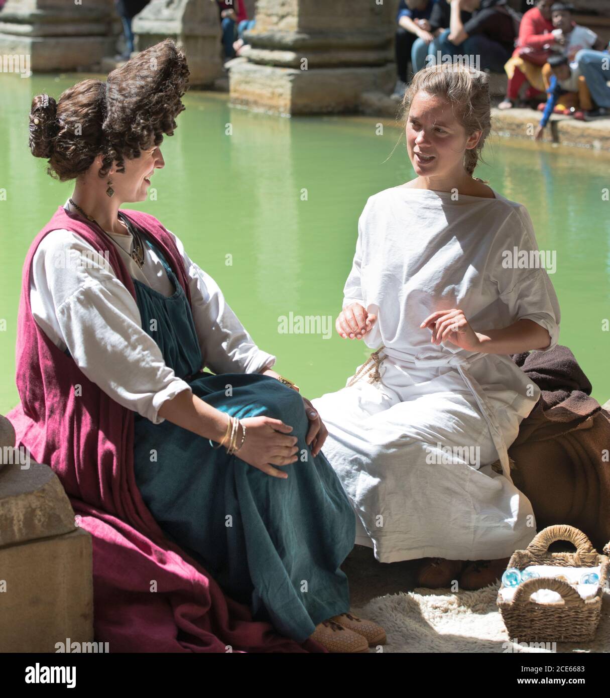 Due donne in costume romano che siedono sul bordo del grande bagno romano, Bath, Inghilterra. Attori femminili in conversazione animata. Estrema acconciatura. Foto Stock