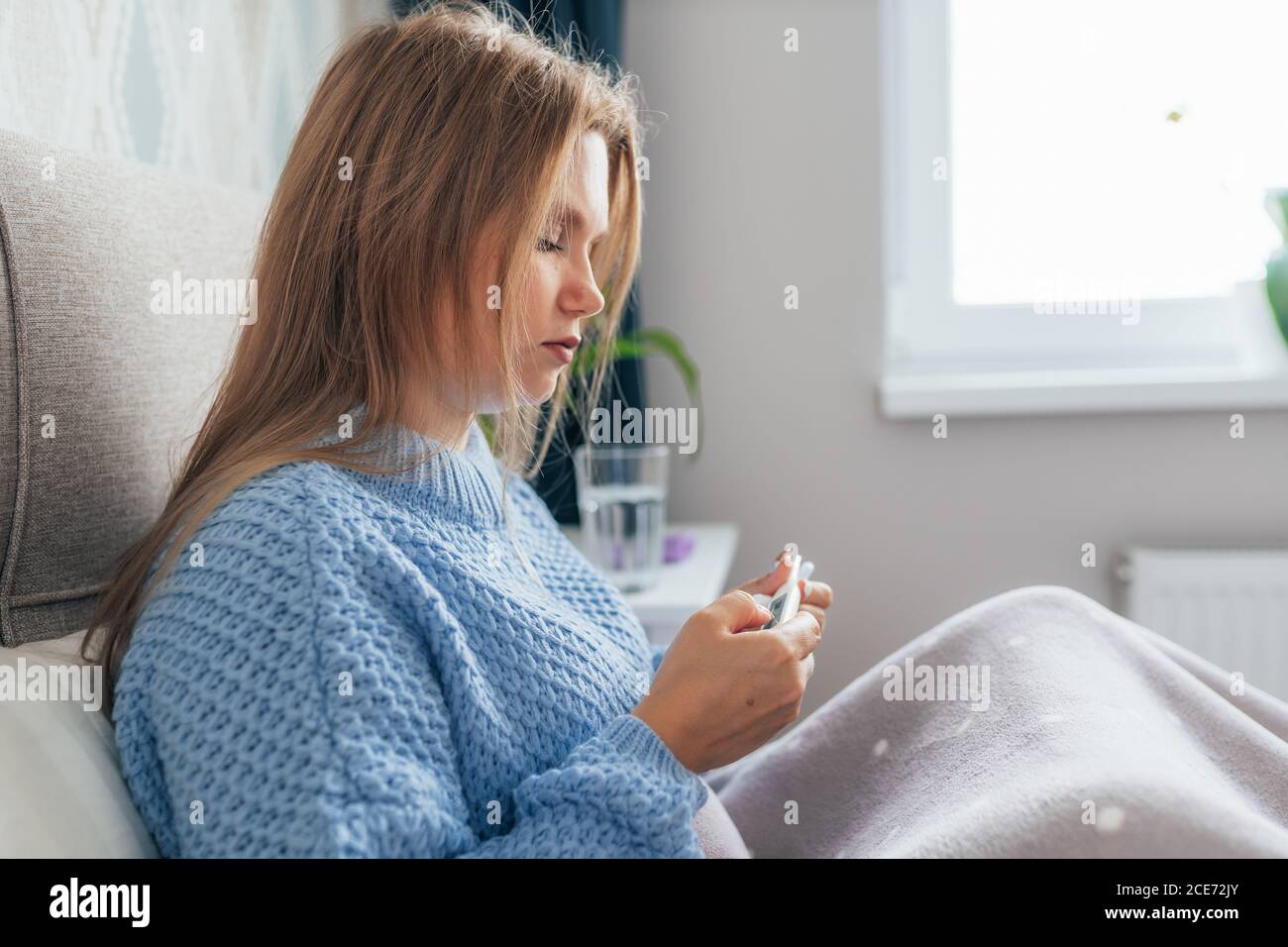 Malata giovane donna bionda si trova a letto a casa sotto coperta e controllare termometro. Mal di testa, influenza, freddo, virus. Foto Stock