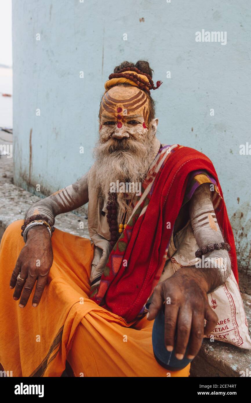 India - 9 marzo 2020: Maschio indiano invecchiato con viso e corpo dipinti e in abiti tradizionali rilassandosi su terra di pietra e guardando la macchina fotografica Foto Stock