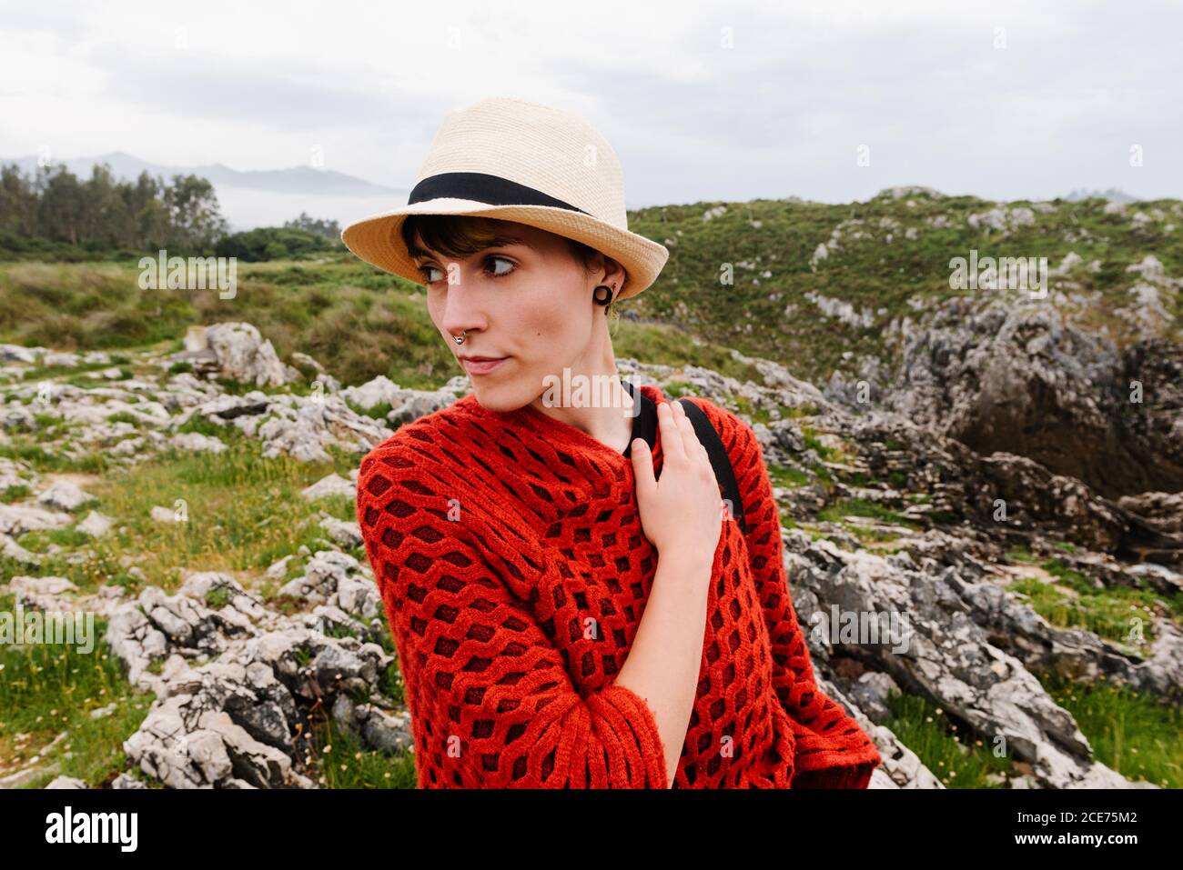 Donna viaggiatore in rosso poncho lavorato a maglia e cappello a piedi Collina erbosa mentre godendo le vacanze nelle Asturie in Spagna Foto Stock