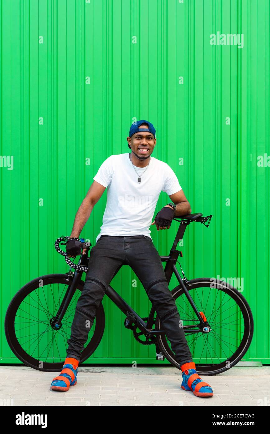 Corpo pieno di allegro giovane afroamericano uomo in casual abbigliamento  street style e scarpe alla moda in bicicletta parete verde sulla strada e  guardando via Foto stock - Alamy