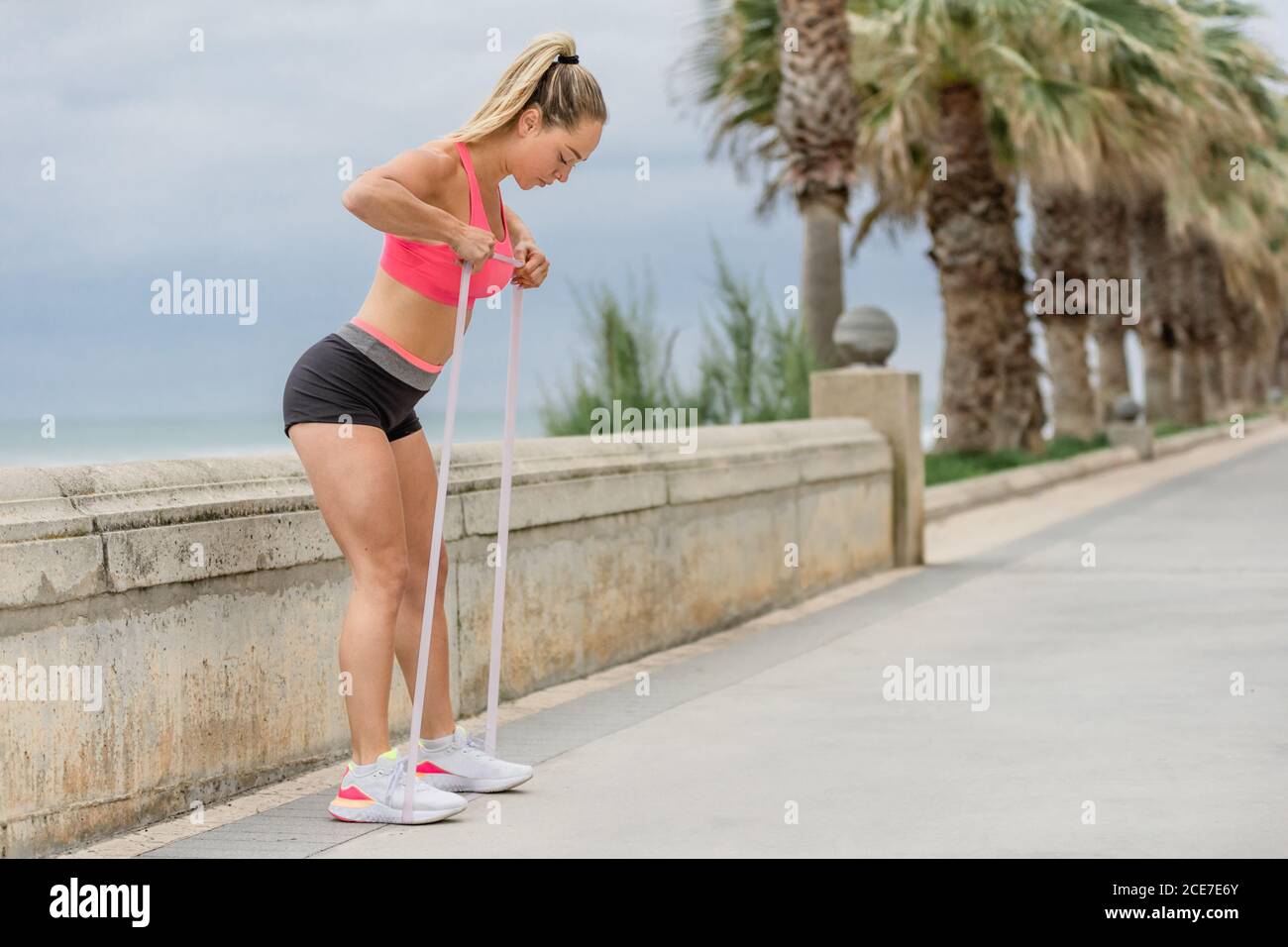Vista laterale dell'atleta concentrata in abbigliamento sportivo che pratica il fitness con nastro elastico mentre si è in piedi appoggiati in avanti sul terrapieno vicino palme e mare sotto il cielo nuvoloso Foto Stock