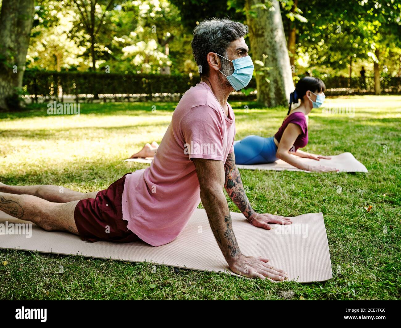 Vista laterale di una coppia serena in maschere protettive che praticano yoga In Bhujangasana su stuoie in verde parco in estate durante focolaio di coronavirus Foto Stock
