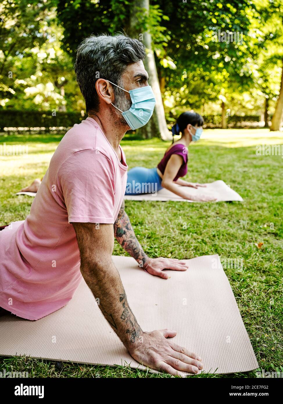 Vista laterale di una coppia serena in maschere protettive che praticano yoga In Bhujangasana su stuoie in verde parco in estate durante focolaio di coronavirus Foto Stock