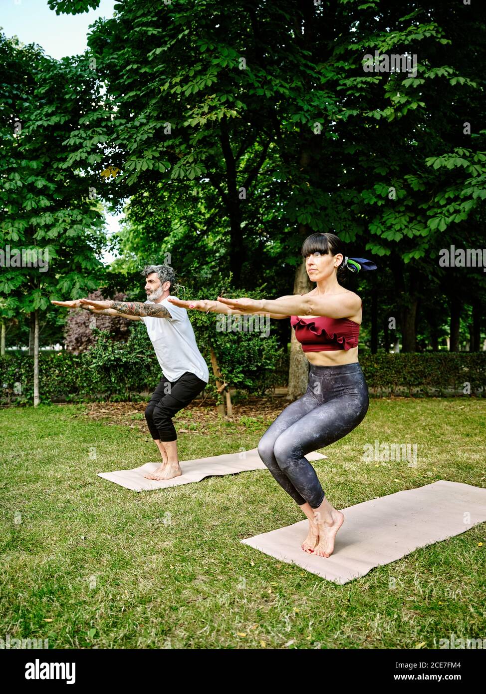 Coppia concentrata in abbigliamento sportivo balancare a piedi nudi su tappetini in Utkatasana mentre si pratica yoga in giardino verde Foto Stock