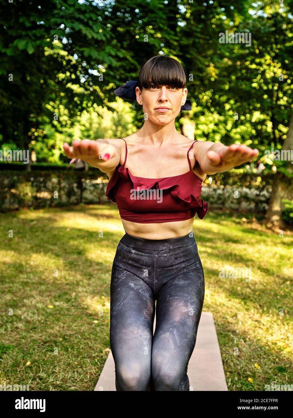 Concentrato sottile femmina in abbigliamento sportivo bilanciamento a piedi nudi su tappetini in Utkatasana mentre si pratica yoga in giardino verde guardando la macchina fotografica Foto Stock