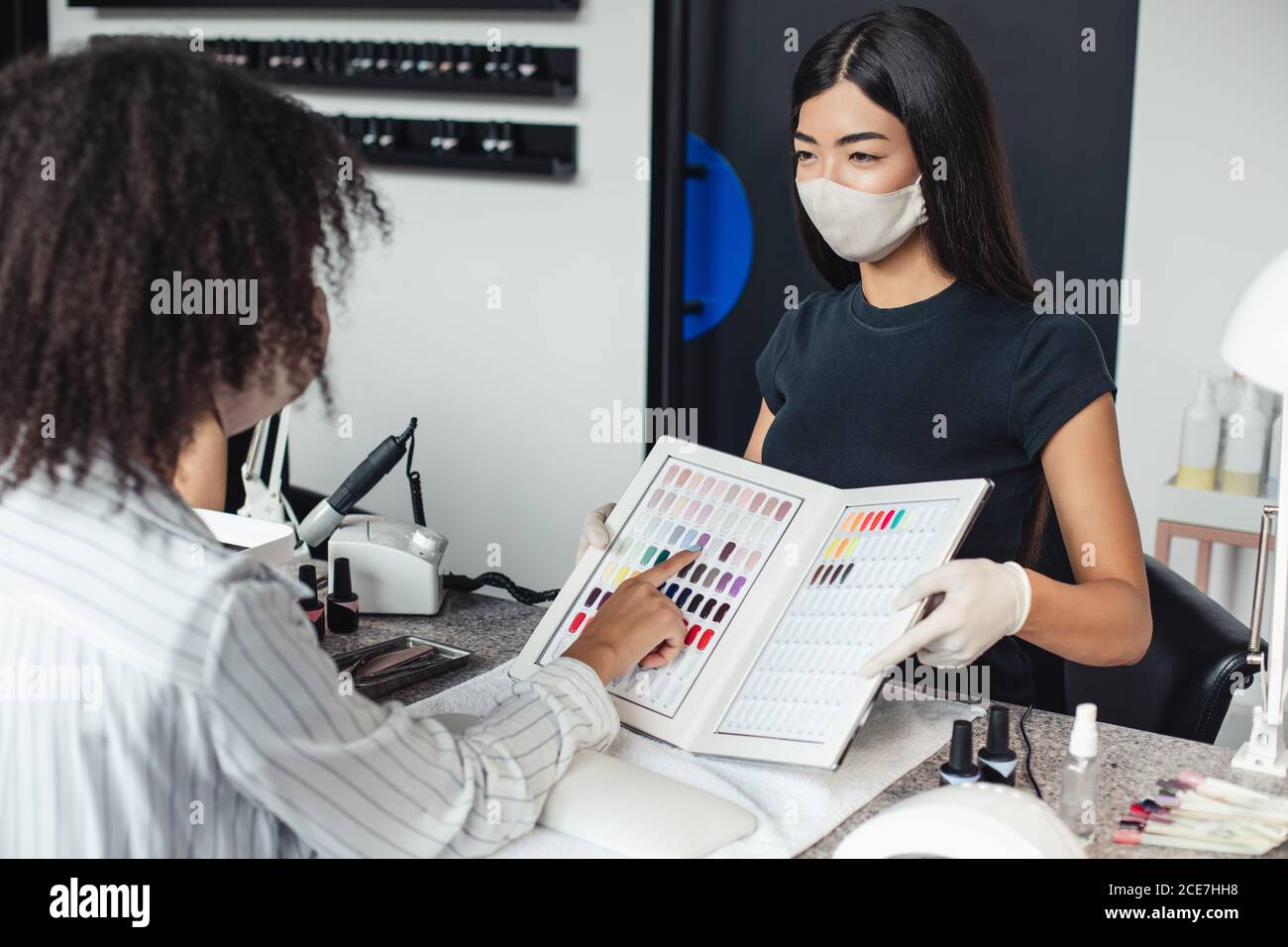 Ragazza asiatica in maschera protettiva guarda afroamericana cliente, sceglie il colore in casa studio unghie Foto Stock