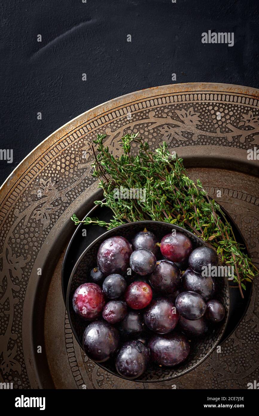 Da sopra ciotola ripiena di deliziose uve rosse mature messe con rametti di timo verde fresco su piatto ornamentale su nero tabella Foto Stock