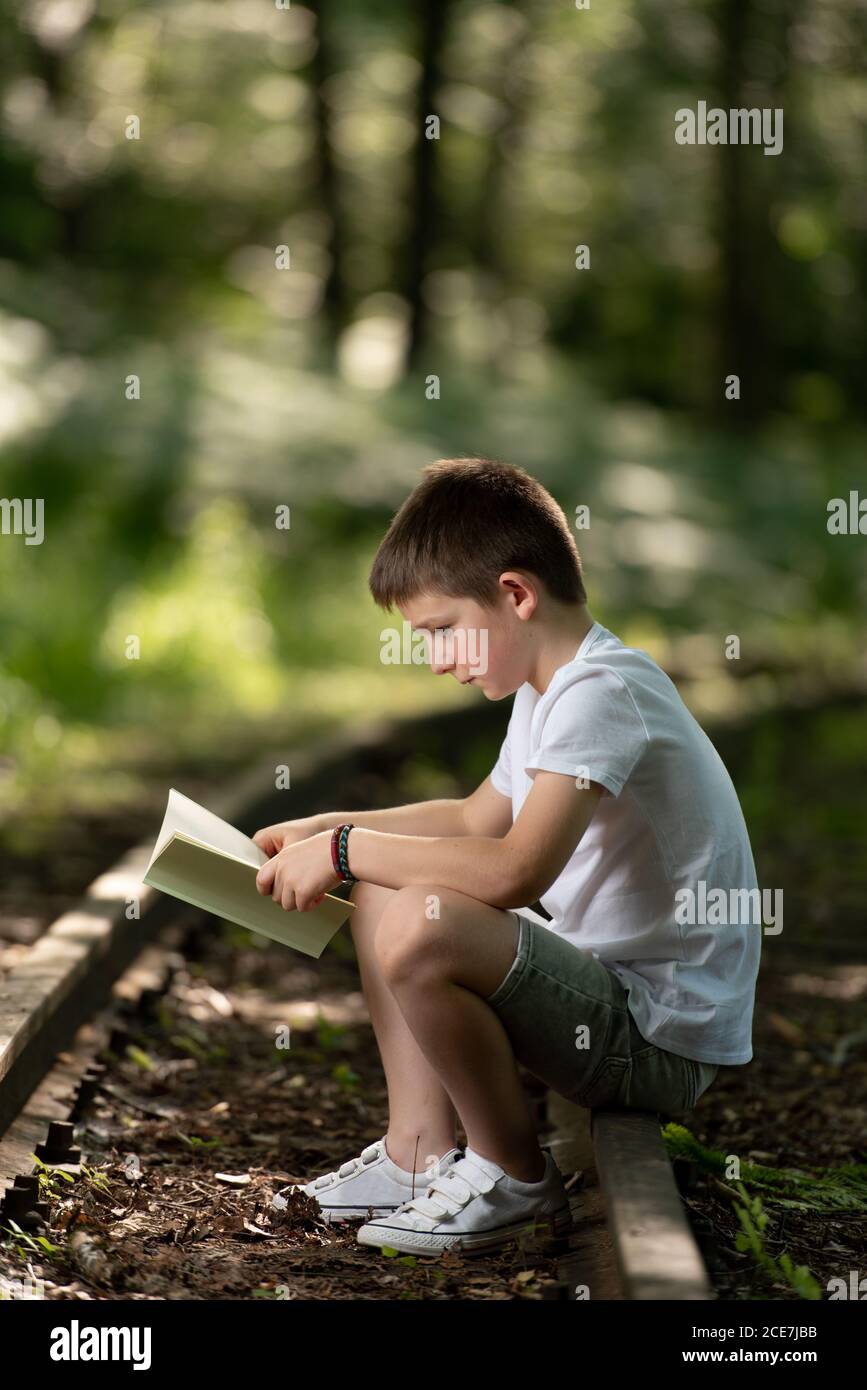 Vista laterale del ragazzo presteen seduto in foresta lettura interessante storia Foto Stock