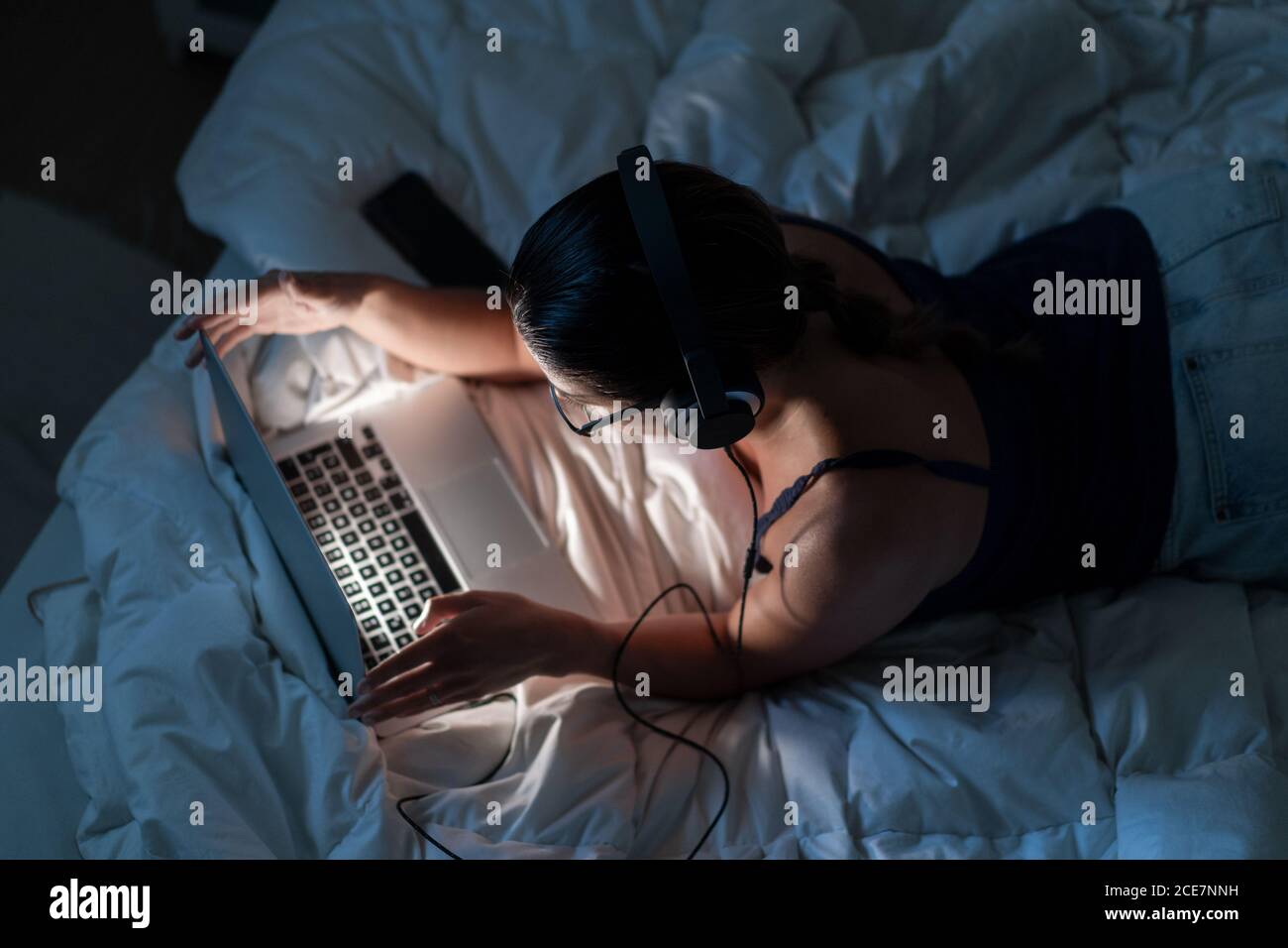 Vista dall'alto sul retro di una femmina rilassata nella parte superiore del serbatoio sdraiato su una coperta sul letto mentre si ascoltano le canzoni laptop in cuffia in serata Foto Stock