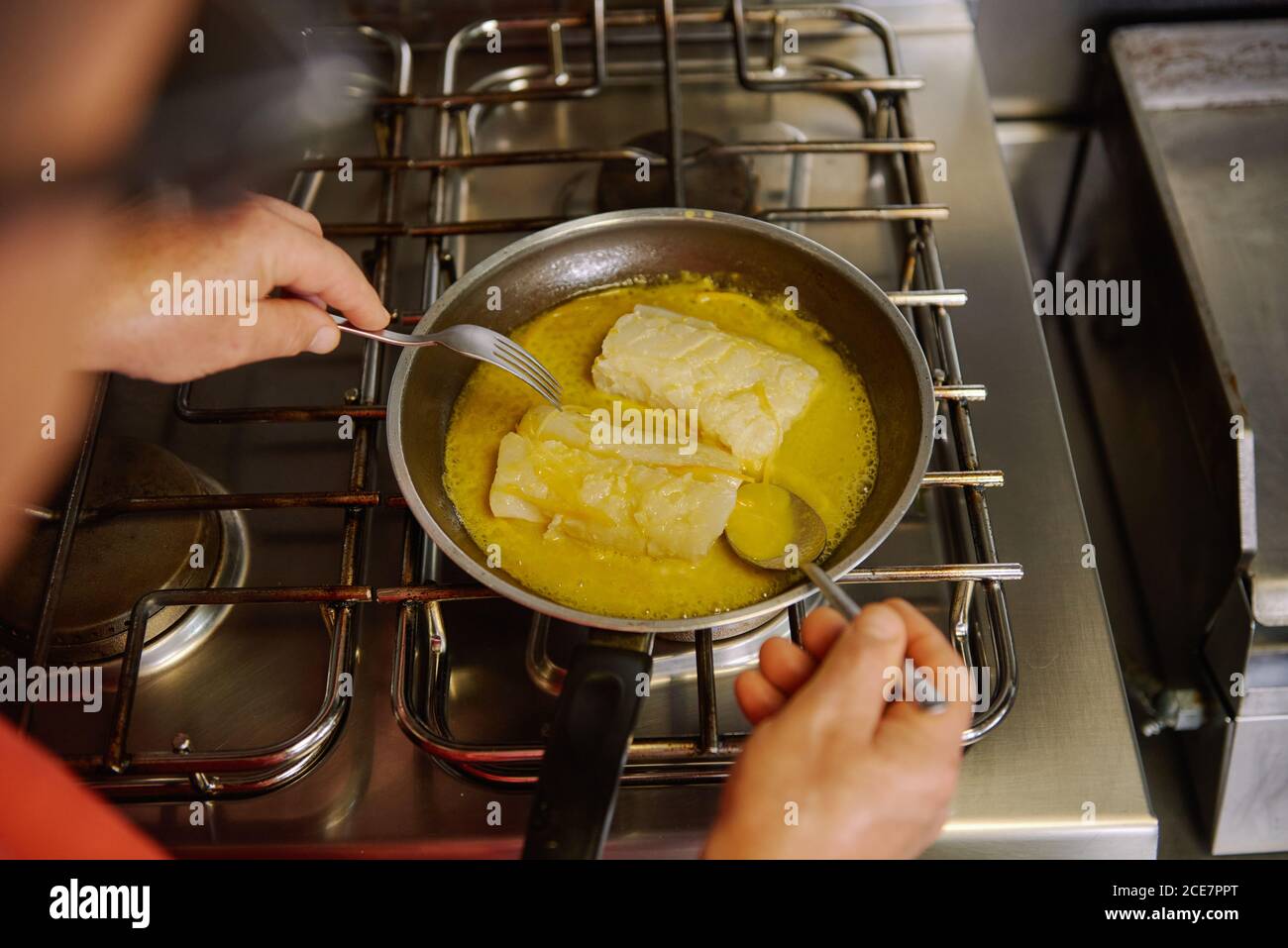 Dall'alto di raccolto unriconoscable chef passando filetti di pesce dentro sciogliere il burro e l'olio d'oliva in padella utilizzando la forchetta e cucchiaio mentre si prepara il cibo a casa Foto Stock