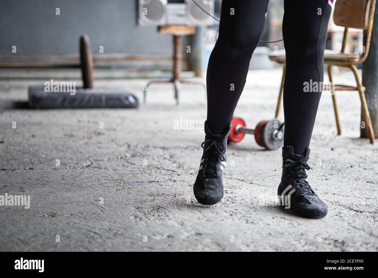 Crop anonymous sportsperson in leggings neri e sneakers che camminano sopra palestra seminterrato con pavimento in cemento vicino a manubri e boombox vecchio stile Foto Stock