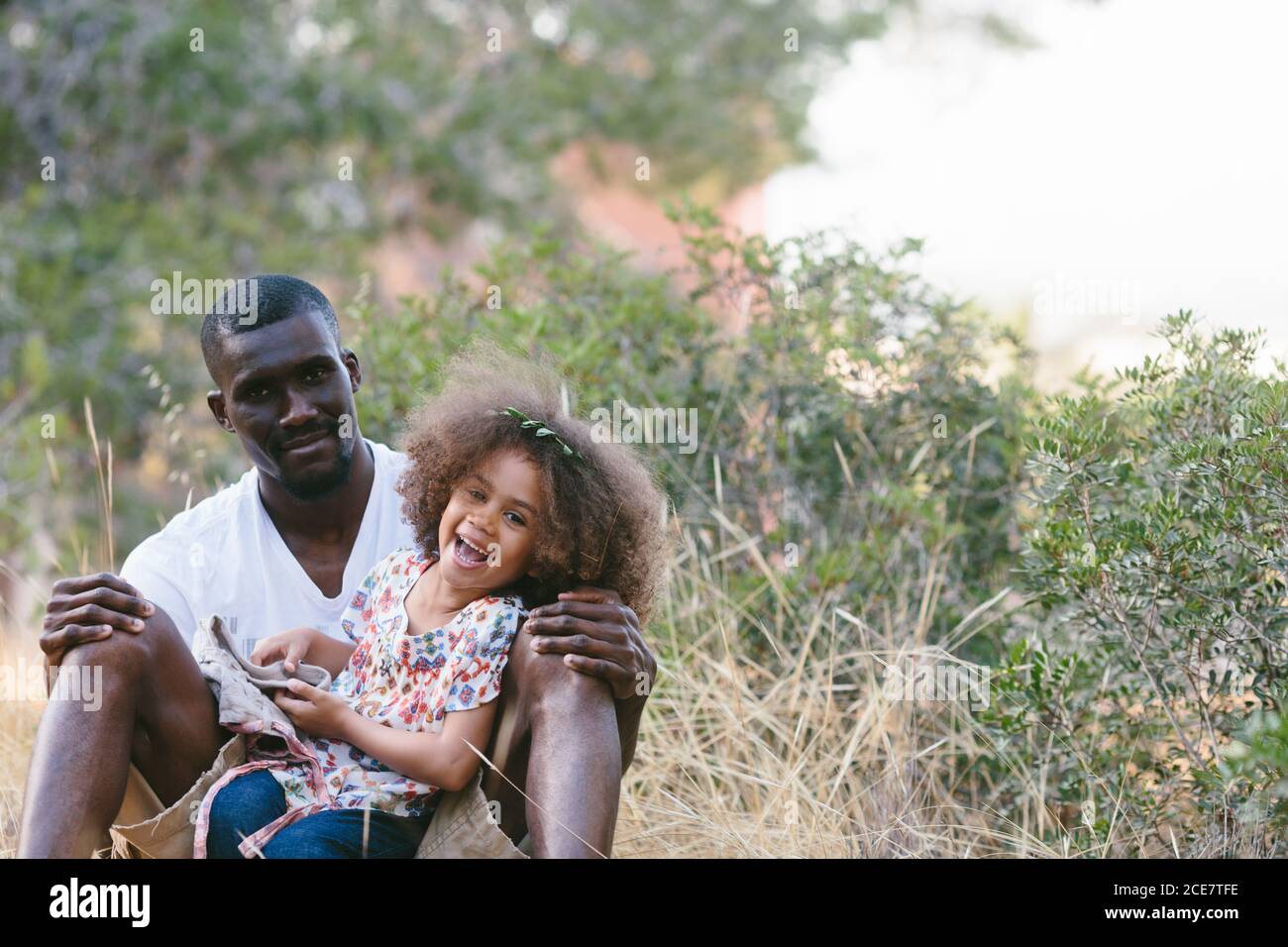 Uomo nero positivo in abiti casual seduto con cute poco figlia sui giri e appoggiandosi sul tronco dell'albero mentre spende giornata estiva soleggiata in foresta Foto Stock