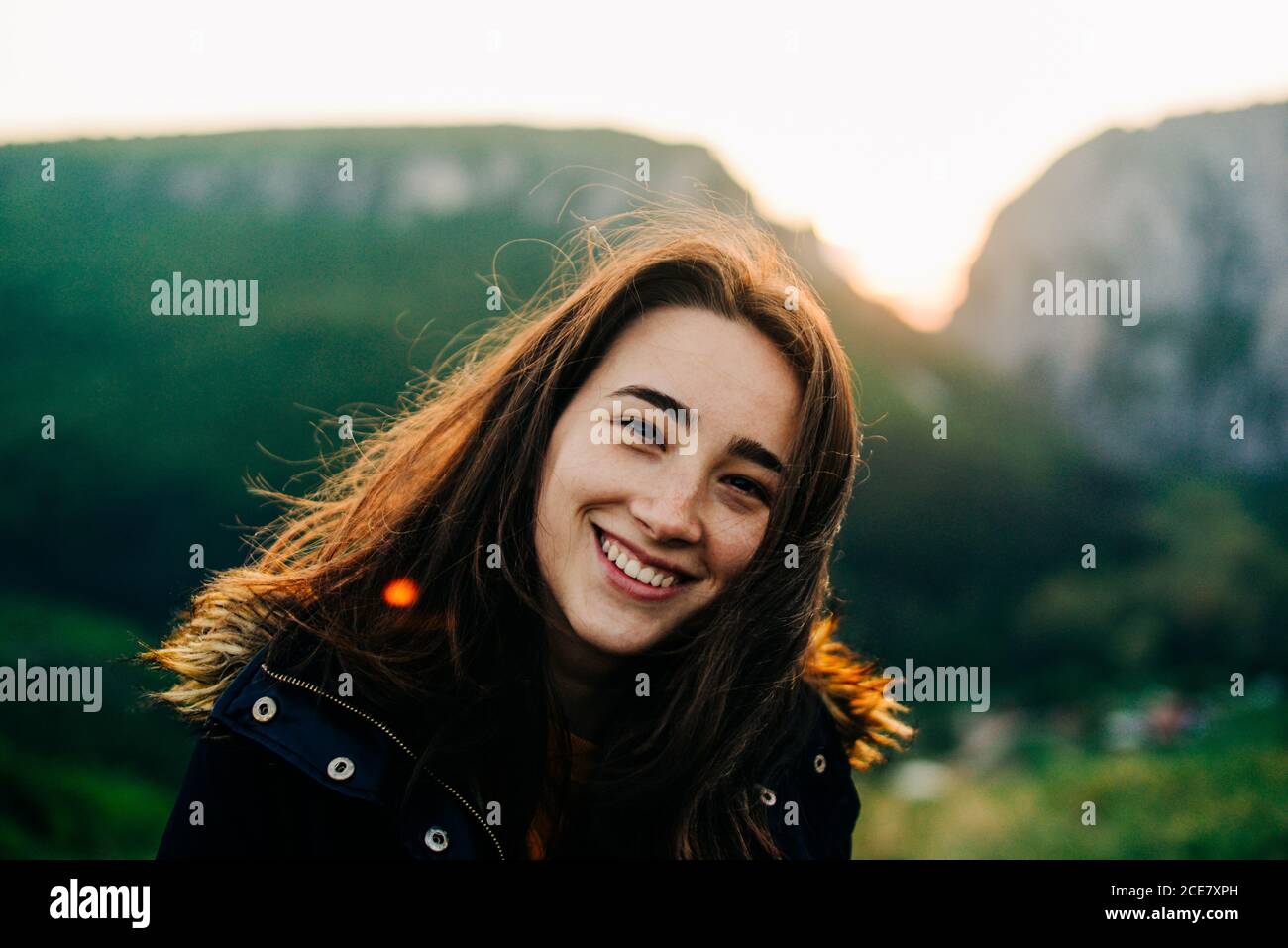 Viaggiatrice femminile spensierata con i capelli volanti che si rilassano nell'area dell'altopiano Al tramonto mentre godendo la vacanza in Transilvania e guardando fotocamera Foto Stock