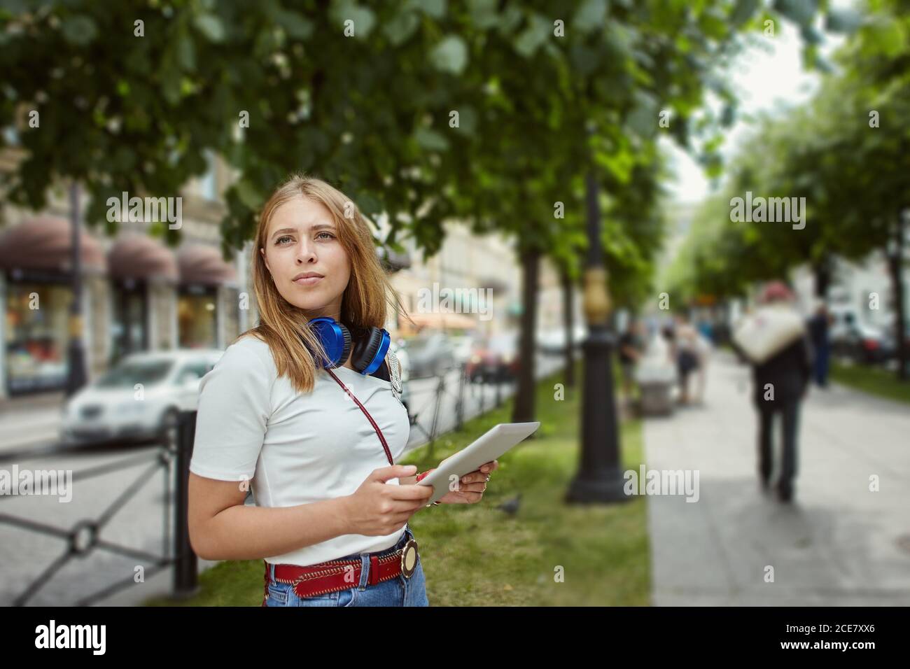 Donna con PC tablet per esterni. Giovane caucasica bella ragazza di circa 20 anni con lunghi capelli biondi e cuffie sta camminando sulla strada della città con Foto Stock