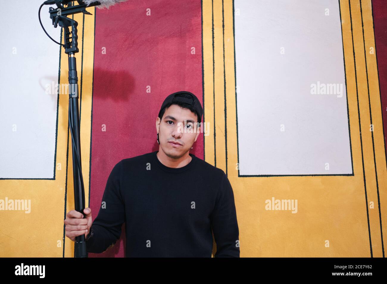 Assistente di videografi focalizzato in camicia nera in piedi con professionista moderno apparecchiature di produzione video su treppiede contro parete creativa in studio Foto Stock