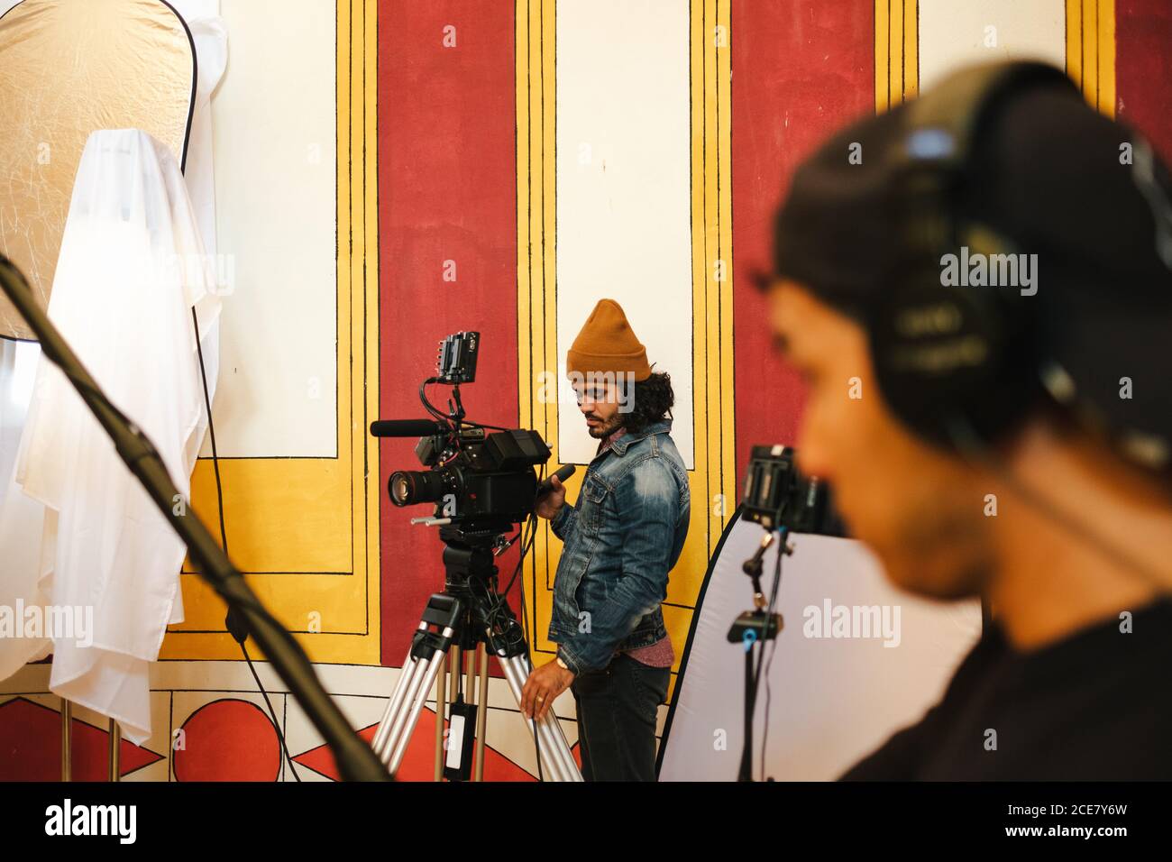 Video operatore serio che riprende video su una fotocamera professionale su treppiede vicino all'assistente con luci Foto Stock