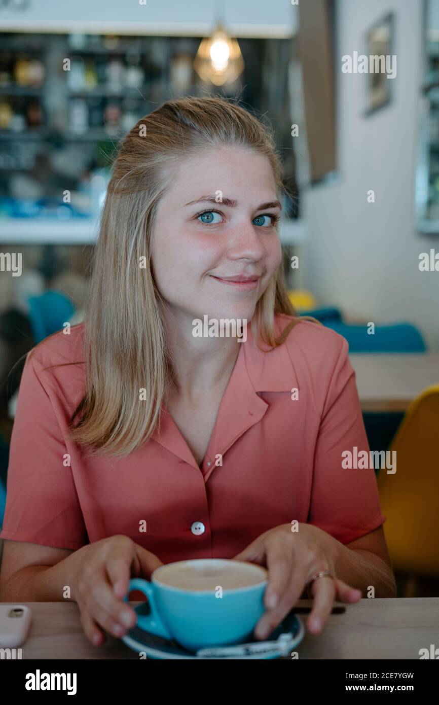 Giovane donna rilassata in abito casual bevendo gustoso tè aromatico da una tazza in ceramica blu mentre si riposa durante la pausa in un ambiente accogliente caffetteria guardando la macchina fotografica Foto Stock