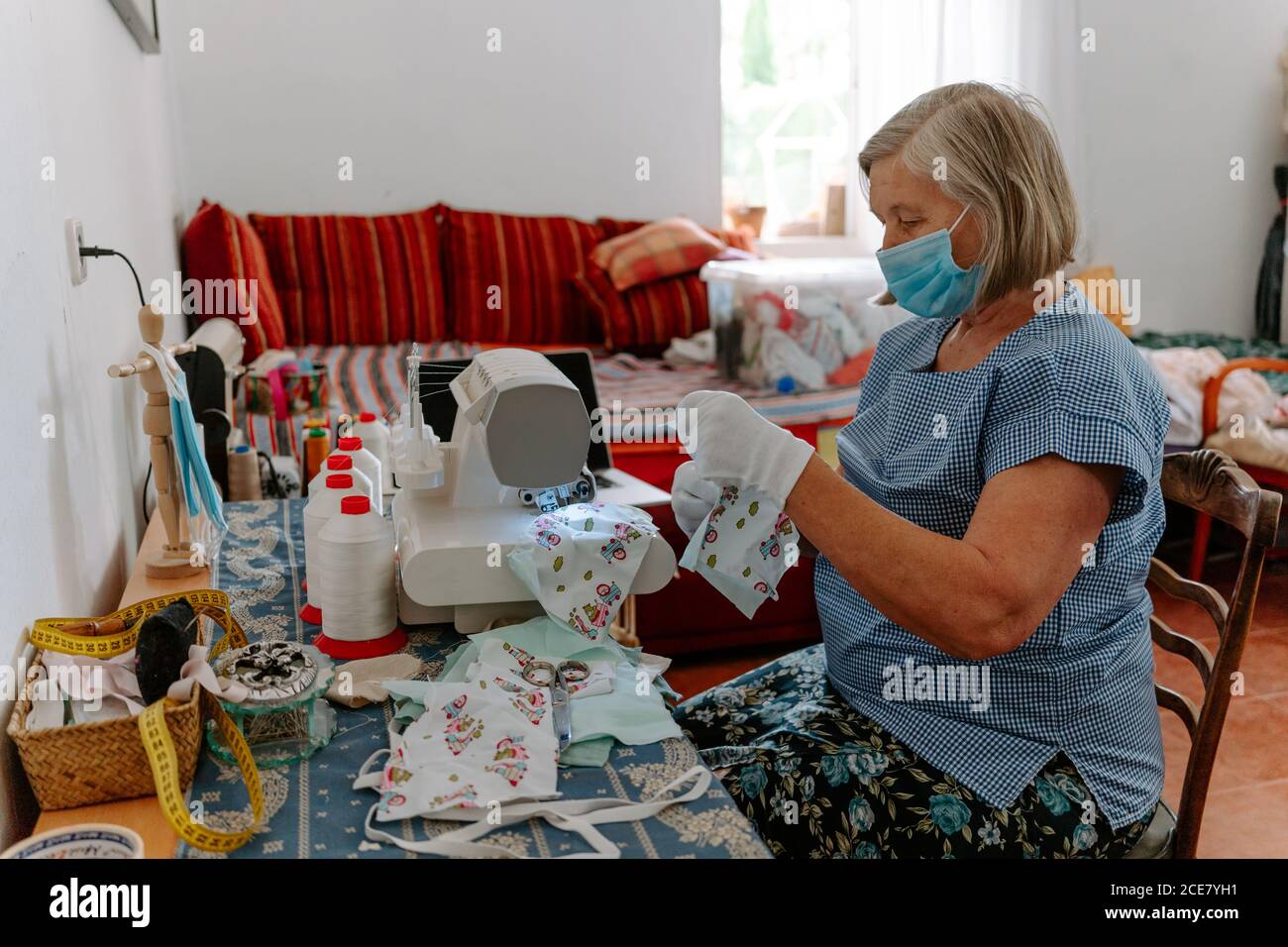 Vista laterale di un'anziana artigiana femminile in maschera medica e.  guanti da cucire maschere per la protezione del coronavirus con tessuto  colorato mentre seduto sul posto di lavoro con macchina da cucire