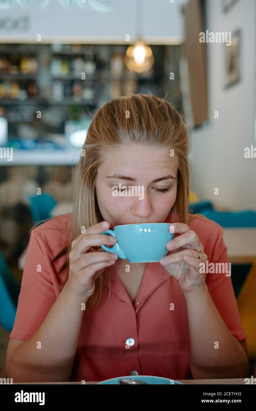Giovane donna rilassata in abito casual bevendo gustoso tè aromatico da una tazza in ceramica blu mentre si riposa durante la pausa in un ambiente accogliente caffetteria Foto Stock