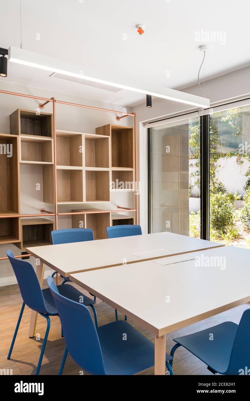 Interni in stile moderno con tavoli quadrati e sgabelli blu su pavimento e ripiani in legno su parete con grande finestra e lampade sul soffitto Foto Stock