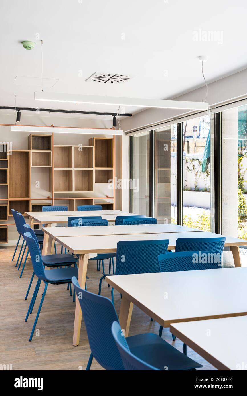 Interni in stile moderno con tavoli quadrati e sgabelli blu su pavimento e ripiani in legno su parete con grande finestra e lampade sul soffitto Foto Stock